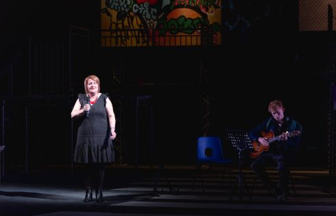 A woman with red hair stands on stage holding a microphone wearing a red necklace and black dress. She is joined by a man sitting down playing guitar. 