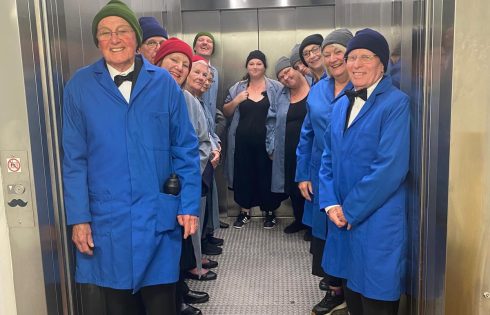 A group of smiling people stand in a lift wearing different coloured beanie hats and blue work overalls. 